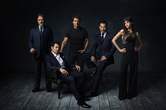 Five actors in formal and dark clothing posing against a dark, textured backdrop.