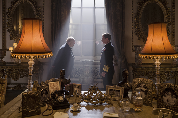 Two men in formal clothing stand in a grand, dimly lit room between two large lamps.