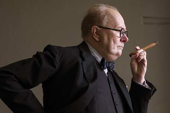 Side profile of an elderly man in a dark suit and bowtie smoking a cigar.