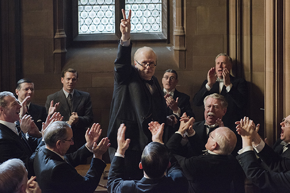 A man in a dark suit stands in a room, holding up two fingers while a group of men clap around him.