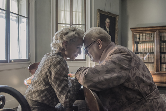 An elderly man and woman sit at a table, leaning their foreheads together in an intimate moment.