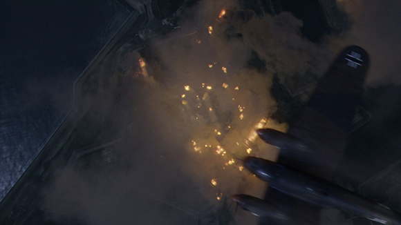 Aerial view of a dark aircraft flying over a coastline with fires and smoke rising from the ground below.
