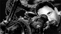 A man peers through the viewfinder of a large, complex professional camera against a dark sky.