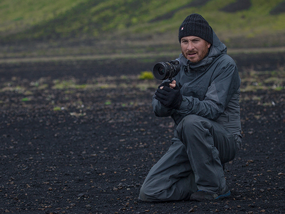 A man in a grey jacket and black beanie kneels on dark volcanic soil, holding a professional camera.