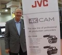 A smiling man in a grey blazer stands beside a large white JVC banner advertising 4K professional camcorders.