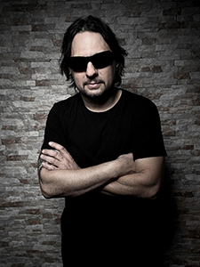 A man with dark hair and sunglasses wearing a black t-shirt, posing with arms crossed against a brick wall.