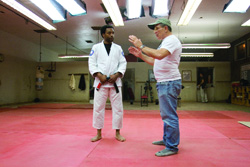 A man in a white gi holds a staff while an instructor in a white t-shirt and jeans demonstrates a movement.