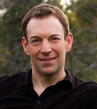A man with short brown hair and a slight smile wearing a black collared shirt.