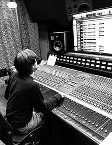 A person sits at a large professional audio mixing console in front of a computer monitor.