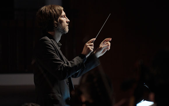 A man in a dark suit conducts with a baton in a dimly lit setting.