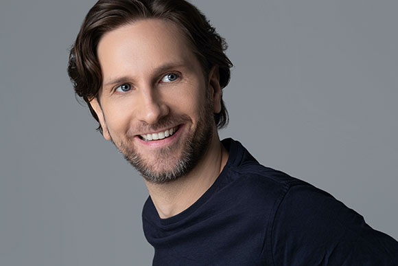 A man with wavy brown hair and a beard smiling while looking off-camera against a gray background.