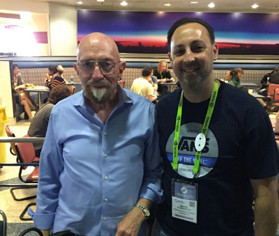 Two men smile for a photo in a convention center. The man on the left wears a blue button-down shirt and glasses. The man on