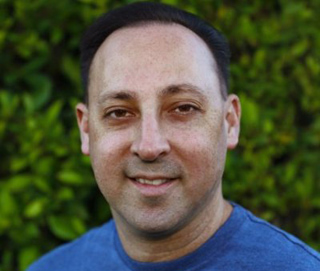 A man with dark hair and a blue shirt smiles.
