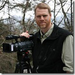 A man with a beard and a vest stands outdoors with a professional video camera on a tripod.