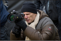 A man wearing a black beanie, scarf, and puffer jacket holds a professional camera to his eye.
