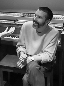 A bearded man in a light sweater sits on a piano bench, looking upward and smiling.