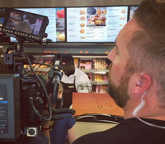 A man with an earpiece watches a camera filming a worker behind a fast-food counter with menu boards above.