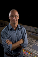 A man with glasses and a blue shirt stands with arms crossed in front of a large sound mixing board.