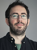 A man with dark hair, glasses, and a beard wearing a black button-down shirt against a gray background.