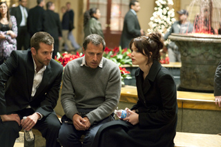 A man in a suit, a man in a grey sweater, and a woman in a black coat sit together on a bench.
