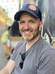 A smiling man with a beard wearing a navy blue cap and a grey t-shirt outdoors.