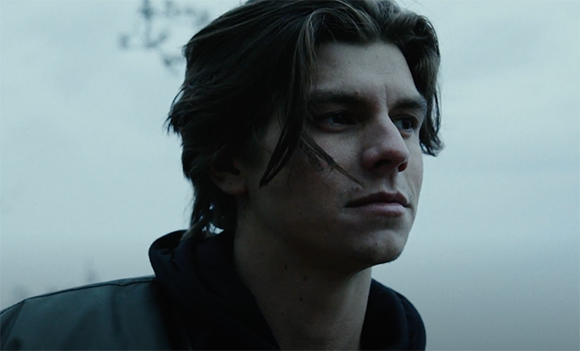 A young man with dark hair looks thoughtfully into the distance under a gray, overcast sky.