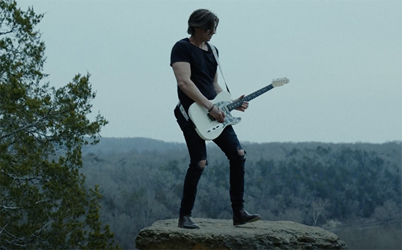A man in black clothing plays a white electric guitar while standing on a large rock overlooking a forest.