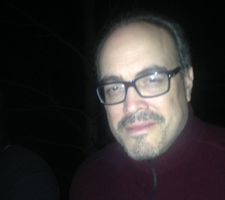 A man with glasses and a goatee looks forward. He is wearing a dark red shirt.