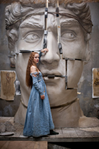 A woman in a long blue dress stands before a massive, cracked stone sculpture of a human face.