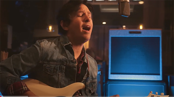 A man in a denim jacket sings passionately into a microphone while playing an electric guitar.