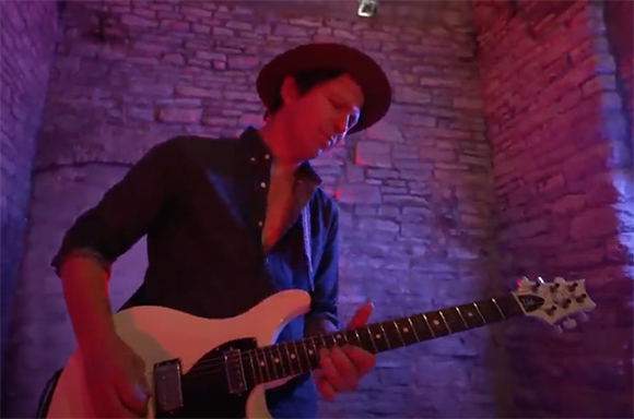 A man wearing a hat and dark shirt plays a white electric guitar under purple lighting.