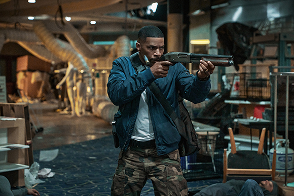 A man in a blue jacket and camouflage pants aims a shotgun in a messy, dimly lit room.