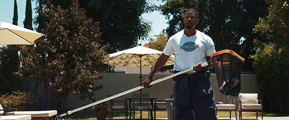 A man in a white t-shirt holds a long pool skimmer pole in a sunny backyard with trees and umbrellas.