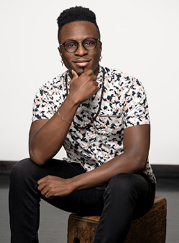 A man with glasses and a patterned shirt sits on a wooden stool, resting his chin on his hand.