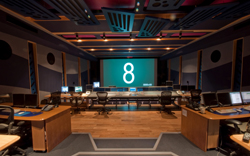 A professional broadcast control room with a large central mixing console and a screen displaying the number 8.