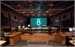 A wide view of a dark news control room with wooden desks and a large teal screen showing the number 8.