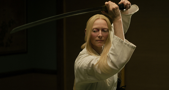 A blonde woman in a white gi holds a katana with both hands above her head.