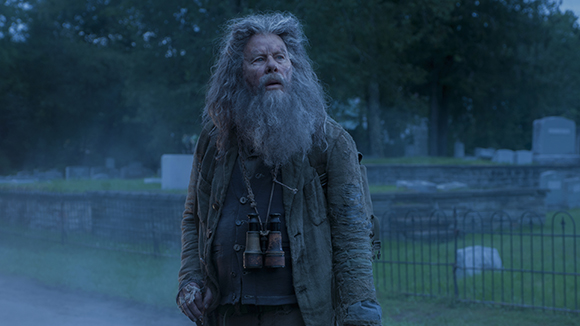 An elderly man with long, messy grey hair and a thick beard stands in a dim cemetery.