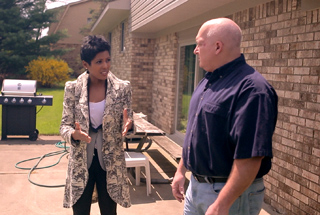A man in a dark shirt and a woman in a patterned coat stand near a brick wall and a grill.