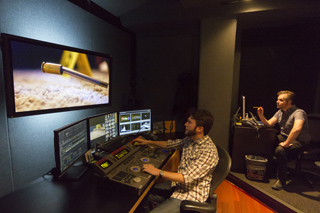 Two men in a dark studio working with multiple computer monitors and a video editing console.