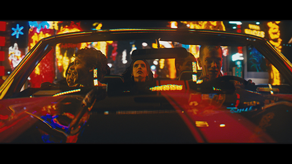 Three people sit in a convertible car at night with colorful city lights blurred in the background.