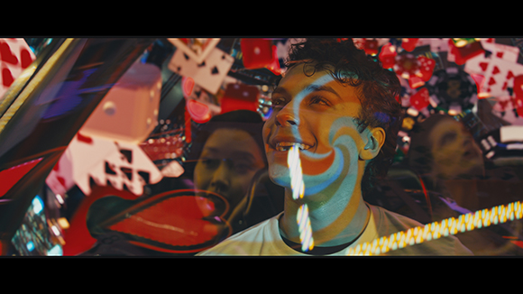 A smiling young man with a blue swirl light pattern projected on his face, surrounded by floating playing cards.