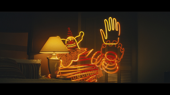 An orange neon sign of a whimsical character with a raised hand sits next to a lamp on a bedside table.
