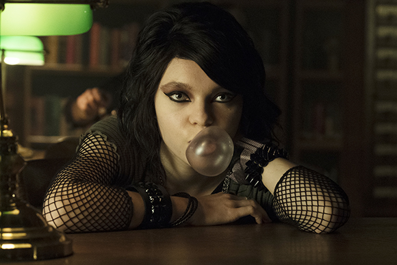 A person with dark hair and fishnet sleeves blows a large bubble with gum while leaning on a table.