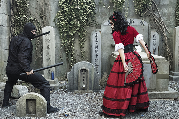 A person in a black ninja suit faces a person in a red Victorian dress among stone graves.
