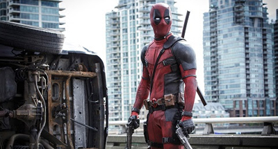 Deadpool in a red and black suit stands next to a wrecked, overturned vehicle in a city.