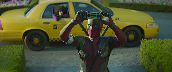 A person in a red Deadpool suit holds up a small cassette player in front of a yellow taxi cab.