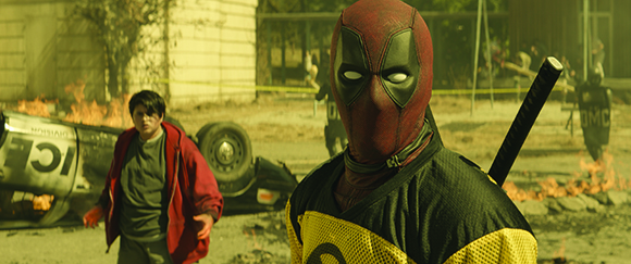 Deadpool in a red mask and yellow jersey stands before a boy and a flipped car in a smoky field.