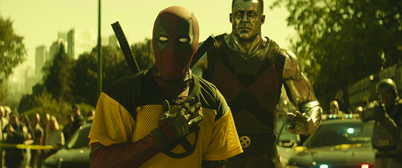 Deadpool in a red and yellow suit stands next to Colossus, a metallic silver character, on a city street.