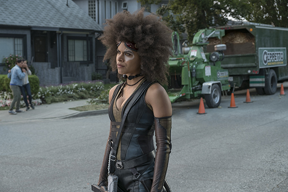 A woman with a large afro and goggles wears a black tactical outfit on a suburban street.